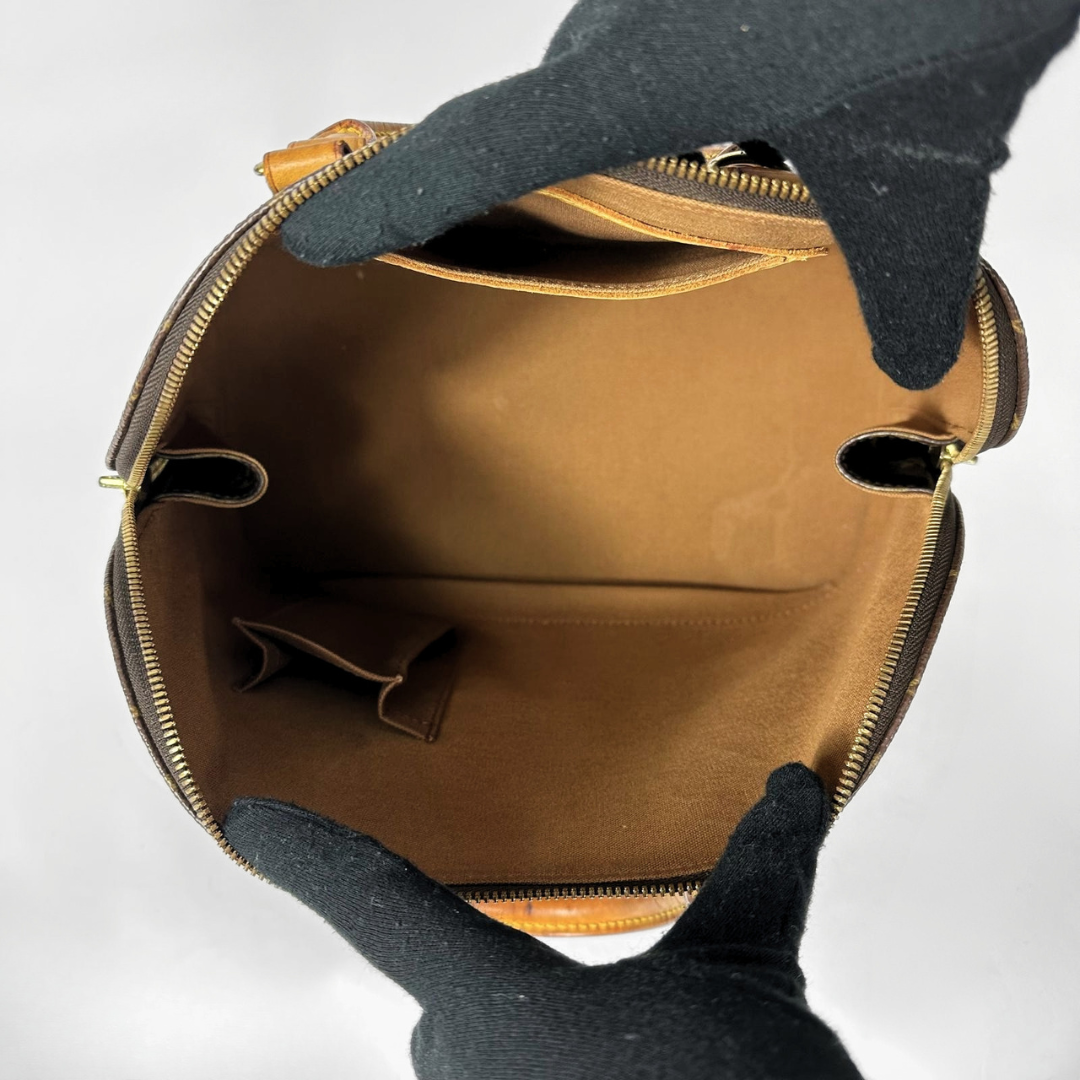 An open brown leather handbag shown from the inside, with a tan interior lining and gold-tone zipper hardware. Gloved hands are holding the bag open against a plain light background.