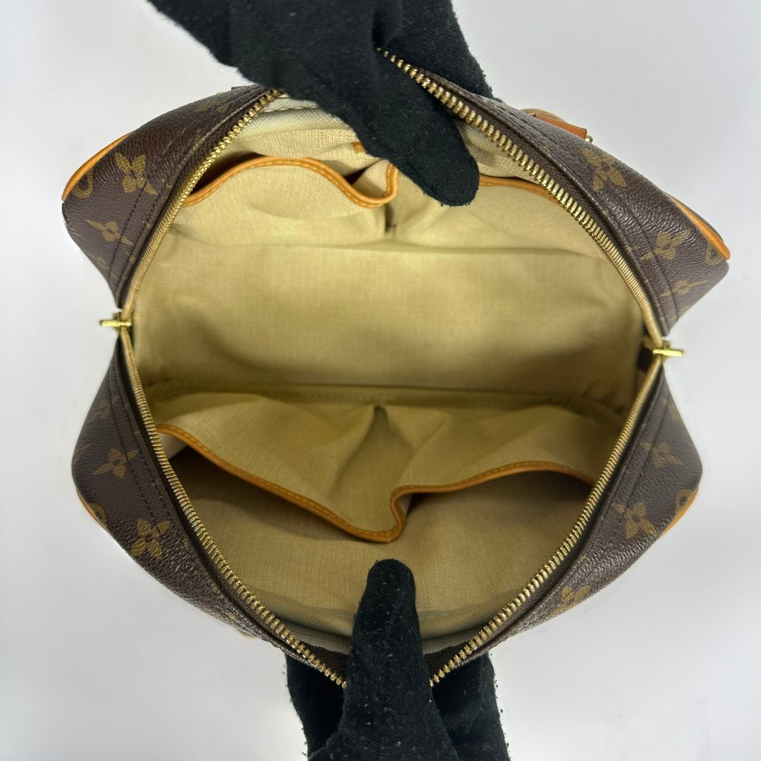 A Louis Vuitton monogram handbag shown open from above, held by black gloved hands against a plain light gray background. The bag has a brown coated canvas exterior with the classic tan monogram pattern, a gold-tone zipper, and a beige fabric interior.