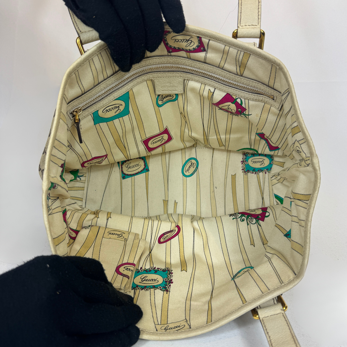 An open beige handbag with a cream interior featuring vertical stripes and small colorful 'Gues' logo patches. Two black gloved hands are holding the bag open, and gold-tone hardware is visible.