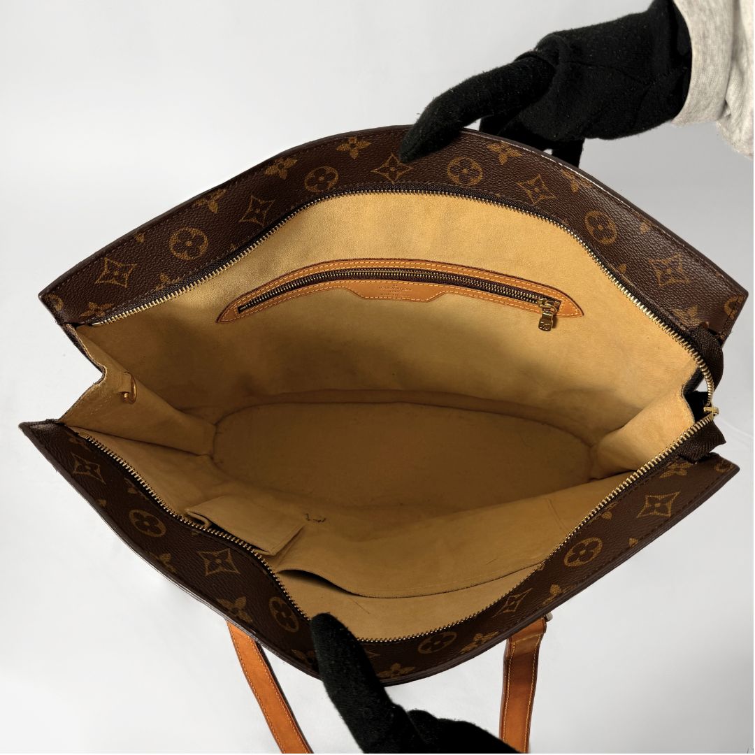 A Louis Vuitton monogram tote bag shown open, with a black-gloved hand holding it against a plain light background. The bag has a brown coated canvas exterior with the classic Louis Vuitton monogram pattern and a tan/beige interior lining.
