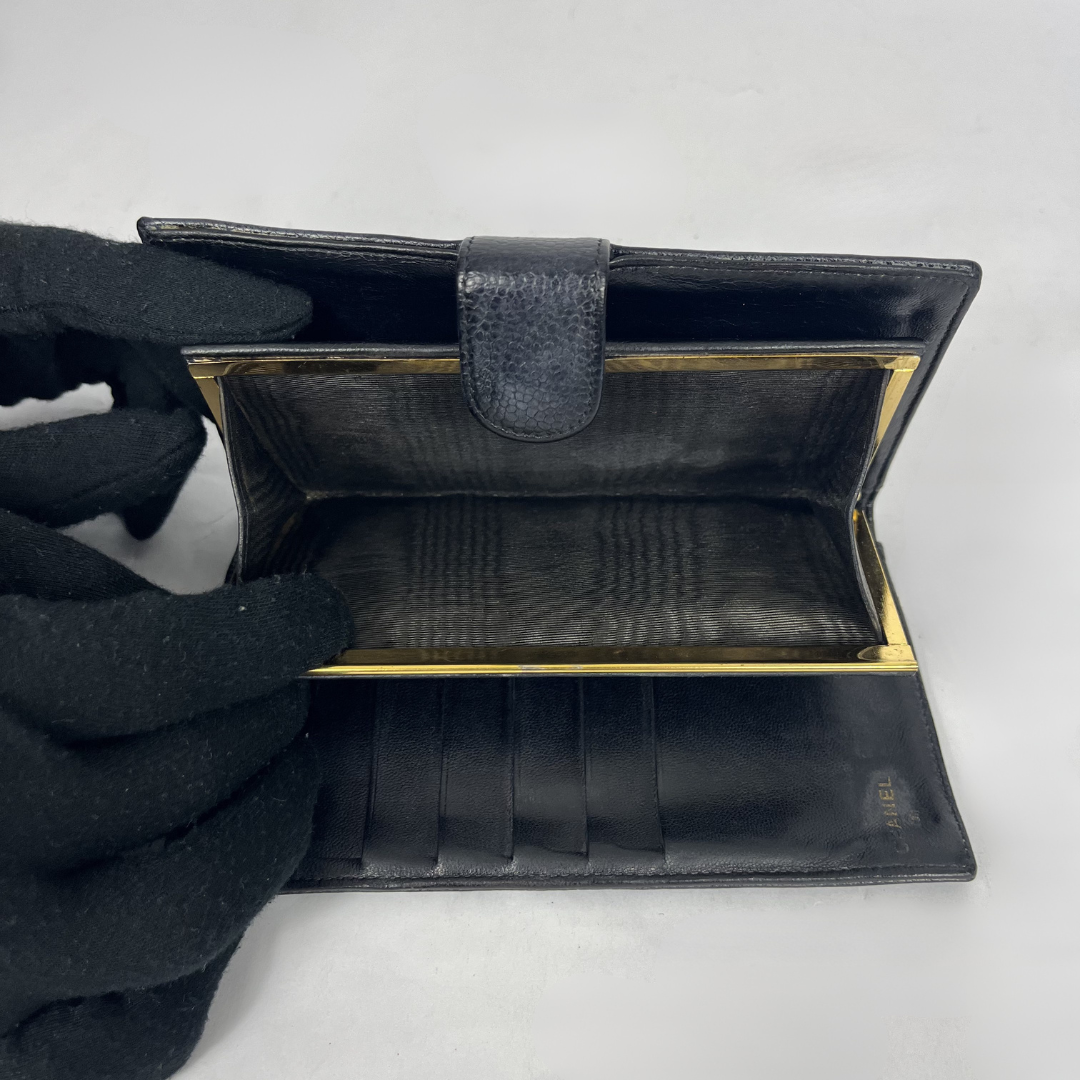 An open black leather wallet with gold-tone metal trim, shown on a light gray background with a gloved hand holding one side.