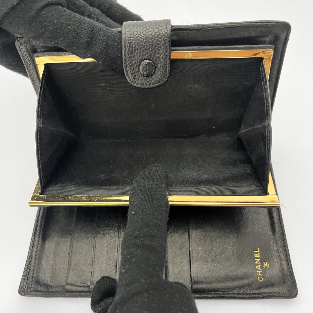 A black leather wallet with gold-tone metal frame details, shown open with gloved hands pointing inside to display the card slots and interior. The Chanel logo is visible in gold on the lower right interior.
