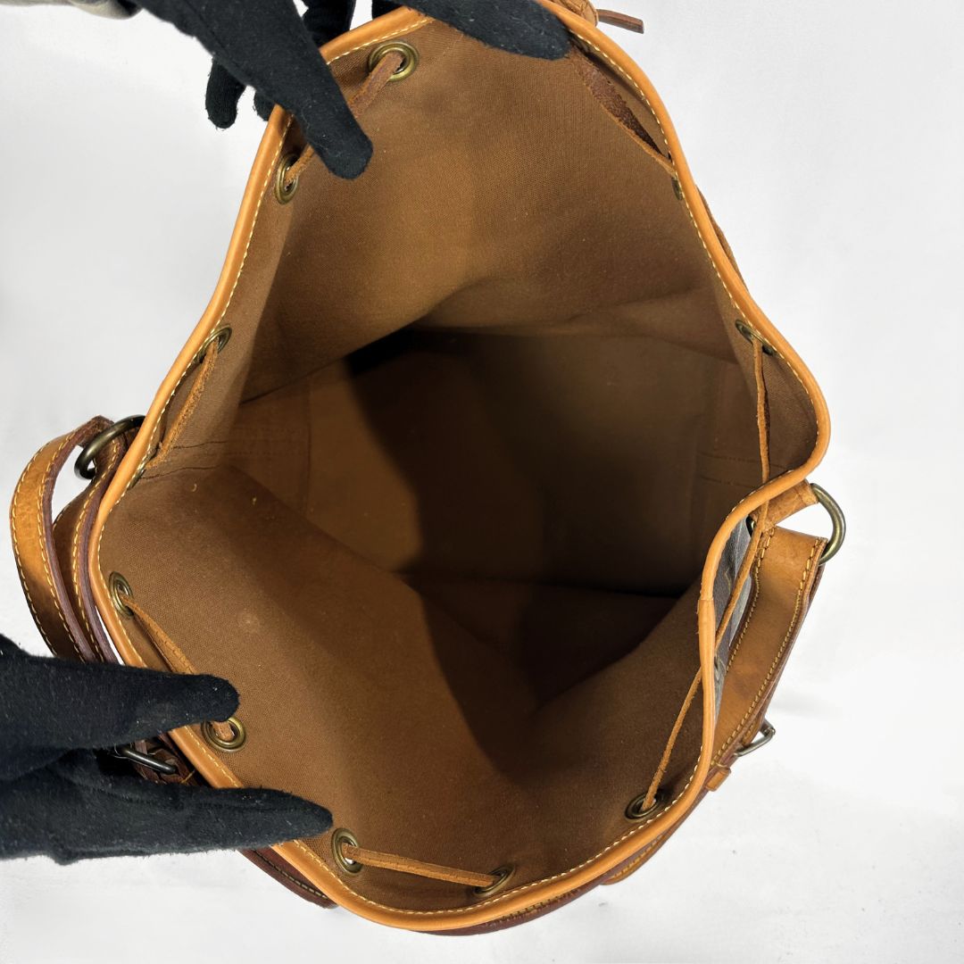 A tan leather bucket bag shown open from above, with black-gloved hands holding the top edge to display the interior. The bag features a tan leather interior, drawstring closure, and brass-tone metal eyelets and hardware.