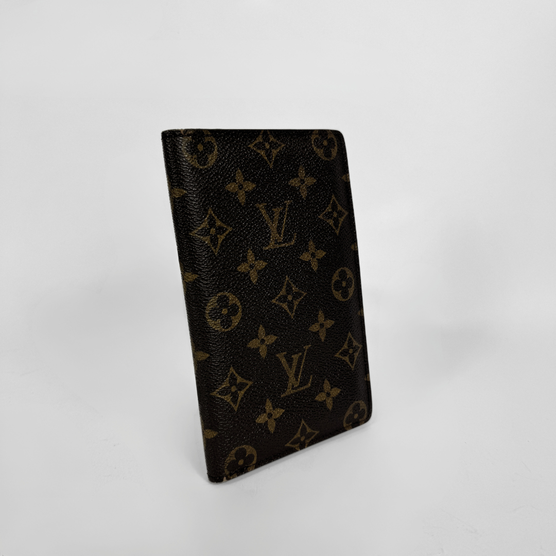 A Louis Vuitton monogram canvas passport cover or document holder in dark brown with tan/gold monogram motifs, standing upright on a light gray background.