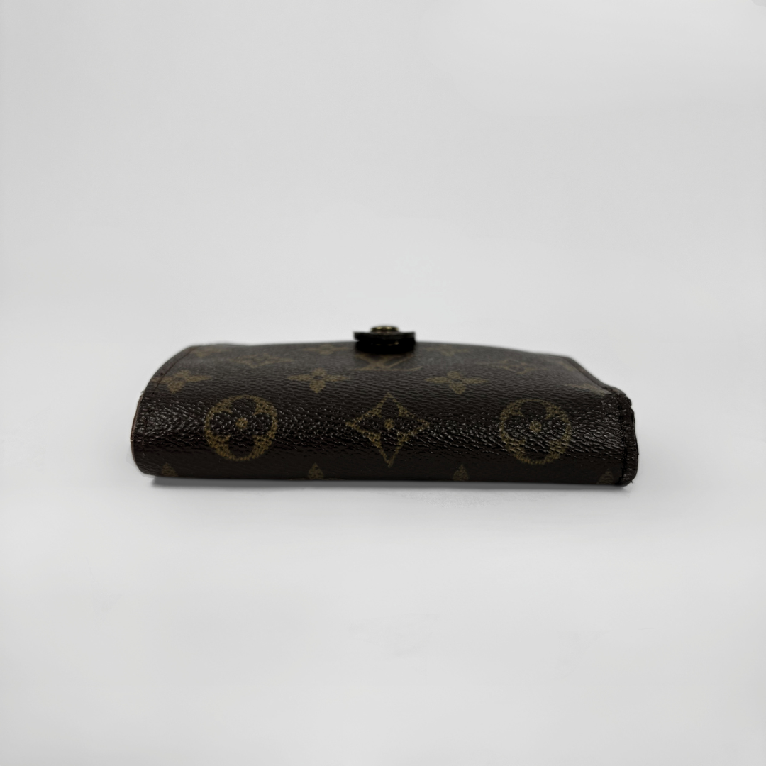 A Louis Vuitton monogram canvas bi-fold wallet in dark brown with gold-tone printed motifs and a small black snap button on top, photographed against a neutral light gray background.