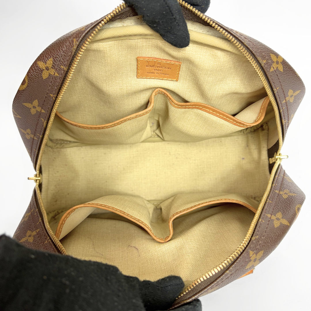 An open Louis Vuitton toiletry bag in brown monogram canvas, showing a beige fabric interior with multiple compartments and gold-tone zipper hardware. Gloved hands are holding the bag open against a plain white background.
