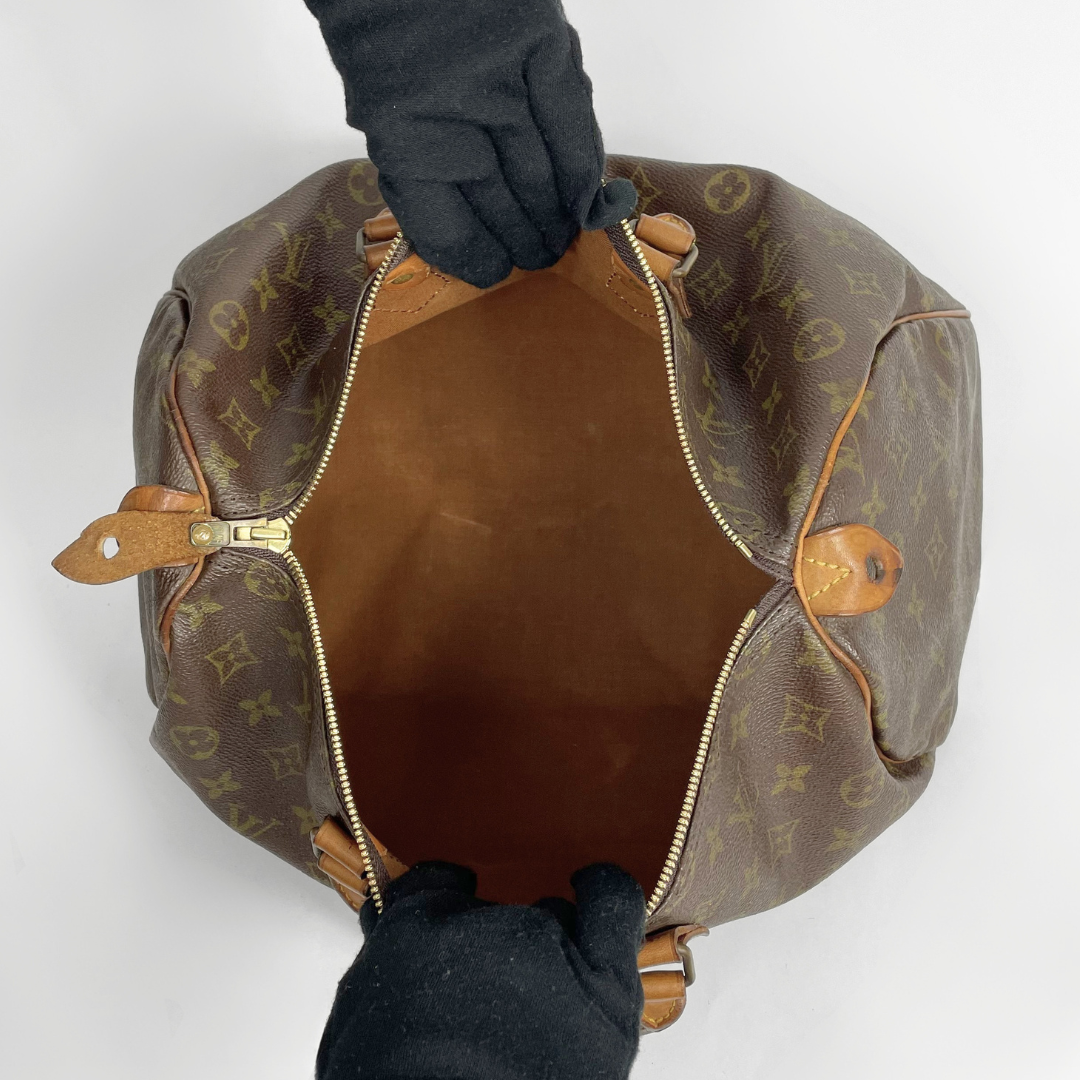 A Louis Vuitton monogram Speedy bag shown from a top-down view, open to reveal the brown interior, with black gloved hands holding it open against a plain white background.