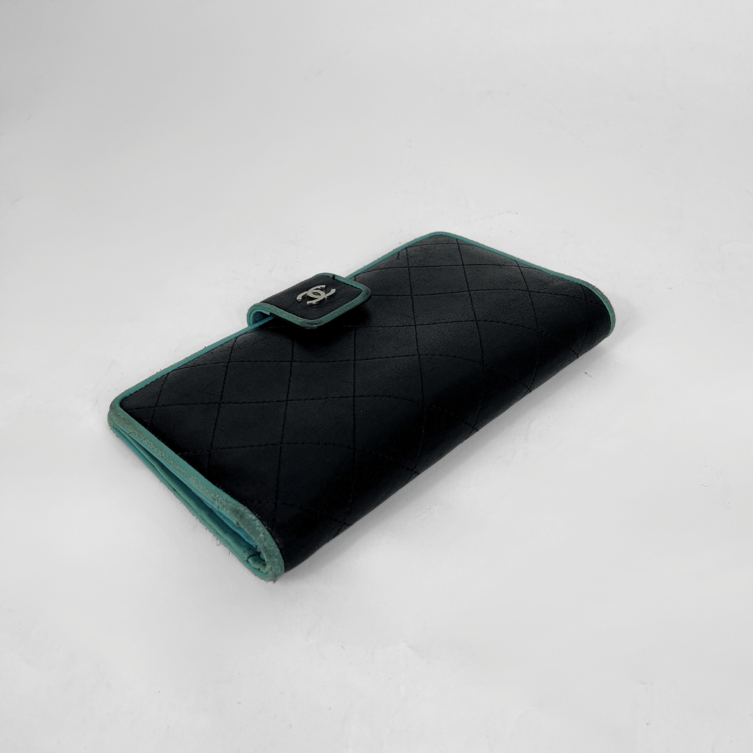 A black quilted leather wallet with teal green trim and a silver-tone interlocking CC logo on the clasp, photographed on a light gray background.
