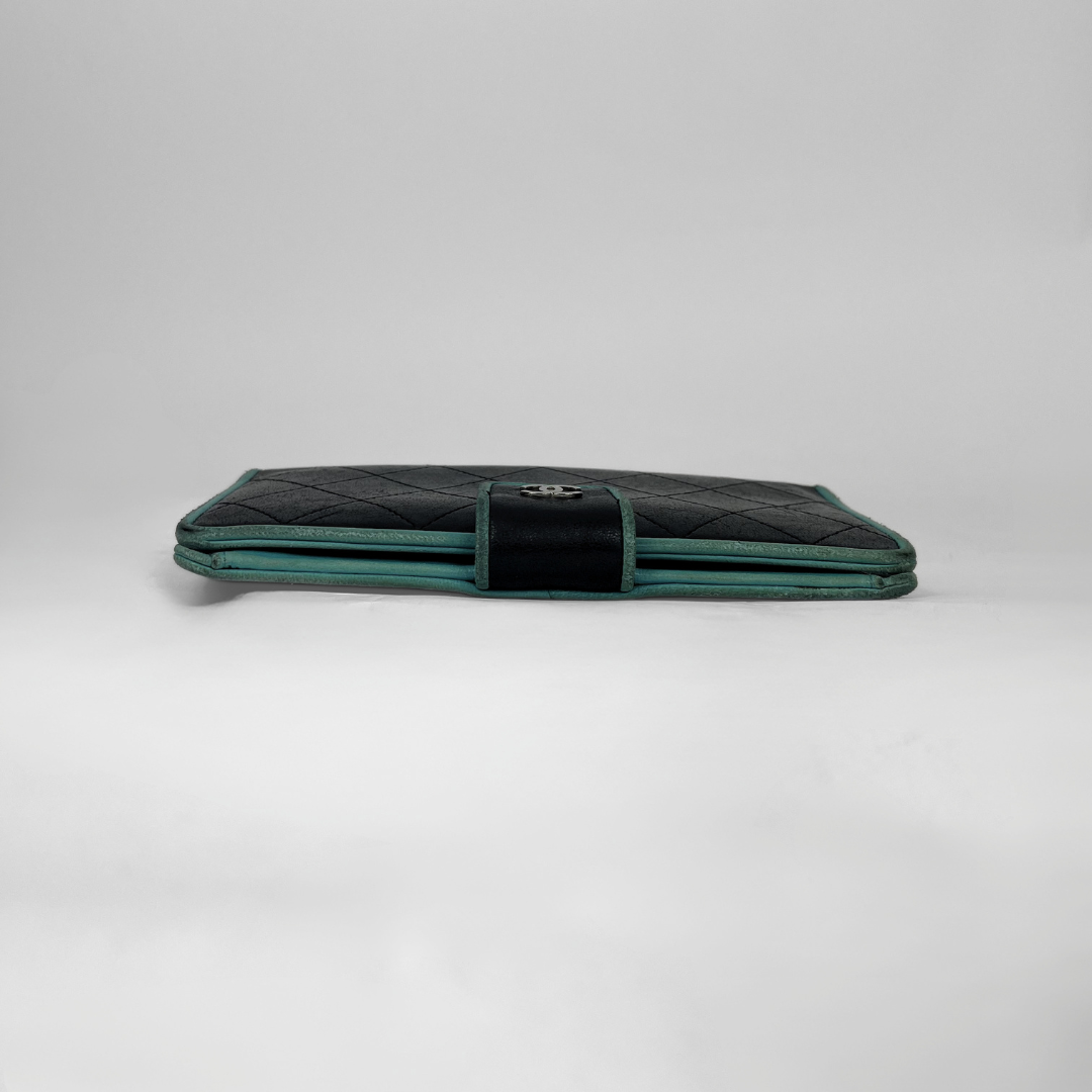 A vintage Chanel long wallet in black quilted leather with a teal green trim and a central black velvet strap featuring a silver-tone interlocking CC logo, set against a plain light gray background.