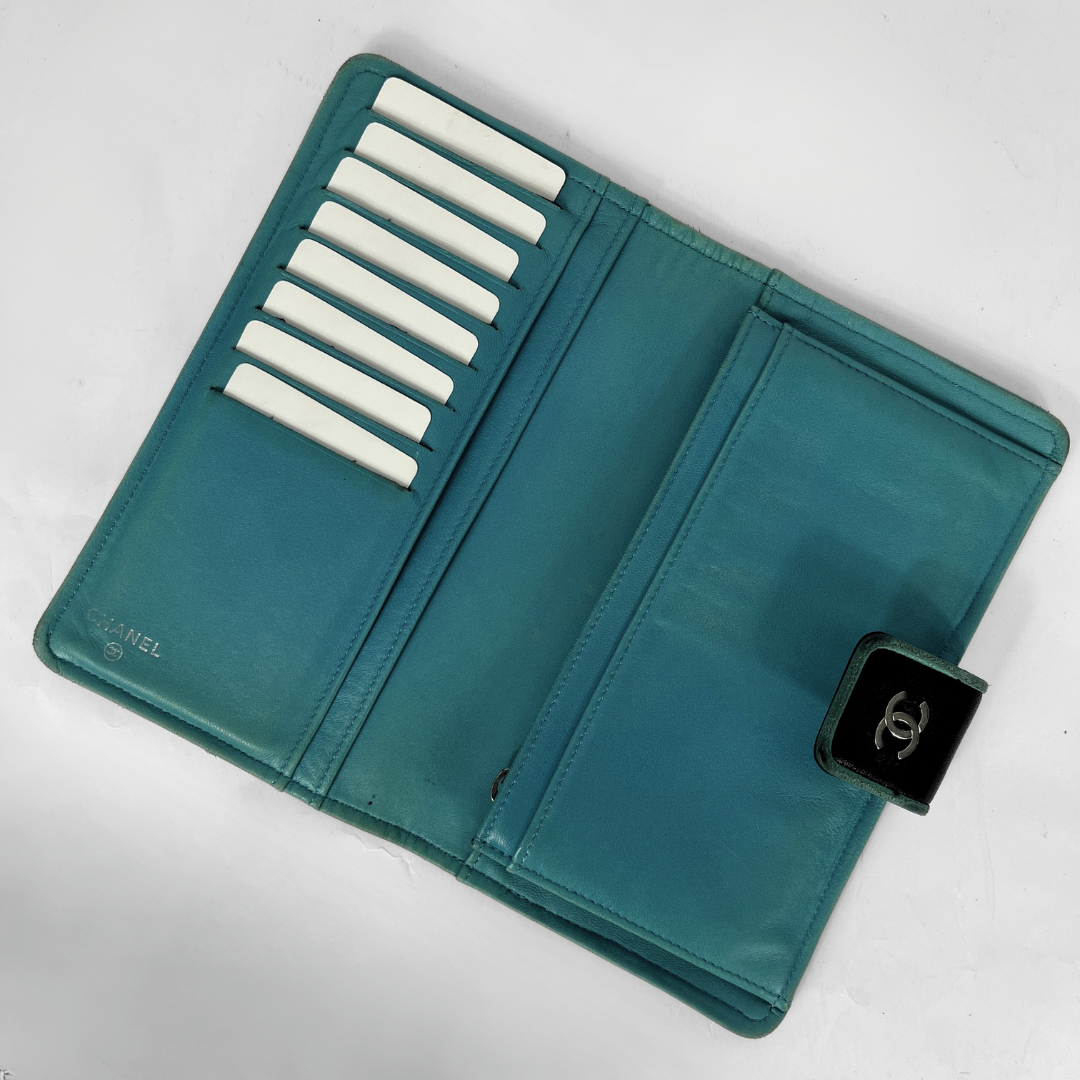 A teal Chanel wallet shown open, featuring multiple white card slots on one side, a large card pocket on the other, and a black leather CC turn-lock closure on the front.