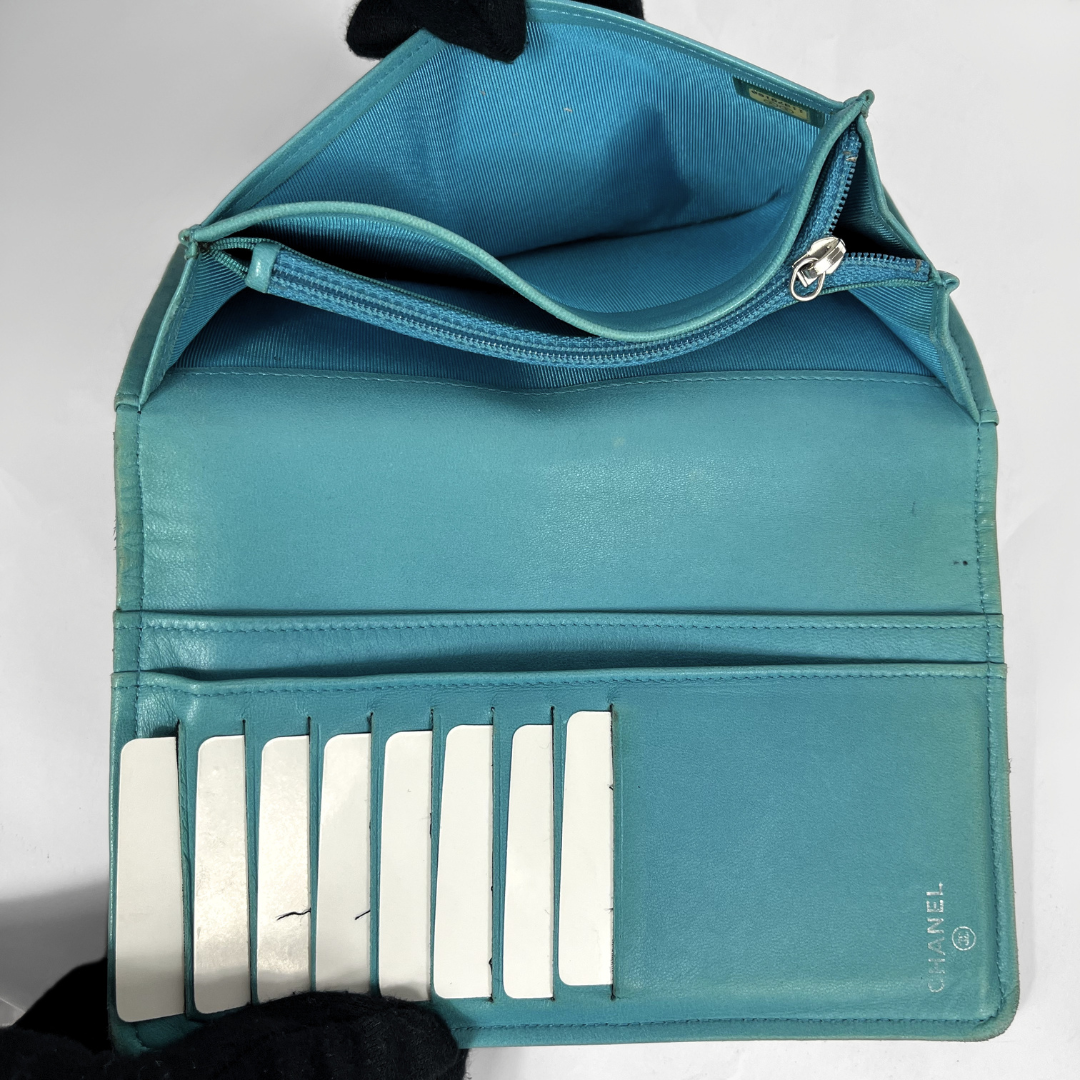 An open teal blue leather wallet or clutch with multiple card slots, a zippered compartment, and a small Chanel logo printed on the lower right corner, shown against a plain light background.
