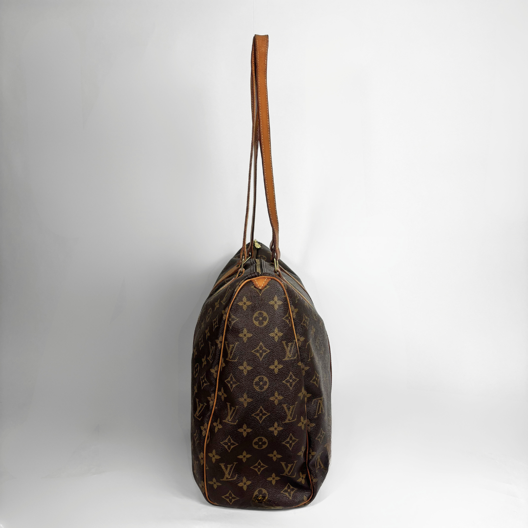 A Louis Vuitton monogram canvas duffel bag with a brown leather shoulder strap, standing upright against a light gray studio background.
