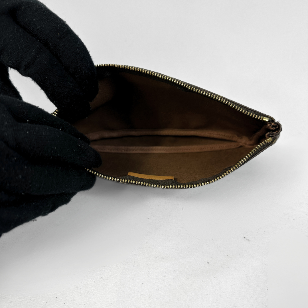 A close-up of a small brown zippered pouch shown open, with a black gloved hand holding one side to demonstrate the interior. The pouch has a dark brown interior lining and a gold-toned zipper.