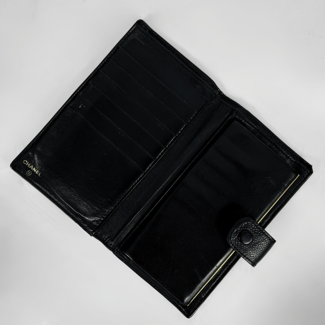 An open black leather wallet with multiple card slots and a coin pocket, featuring a small gold CHANEL logo on the left side, photographed on a light gray background.