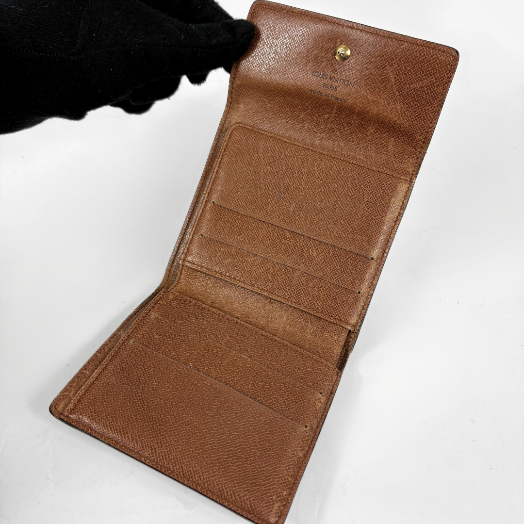 A brown textured leather wallet shown open, revealing multiple card slots and a gold-tone snap button. A black gloved hand is holding the top edge against a plain white background.