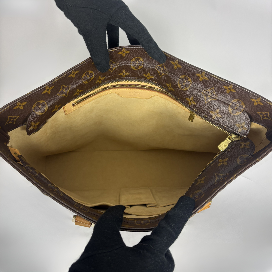A Louis Vuitton monogram handbag shown open, held by black-gloved hands against a plain light background. The bag has a brown coated canvas exterior with the classic tan and yellow monogram pattern, a tan interior lining, and gold-tone hardware.