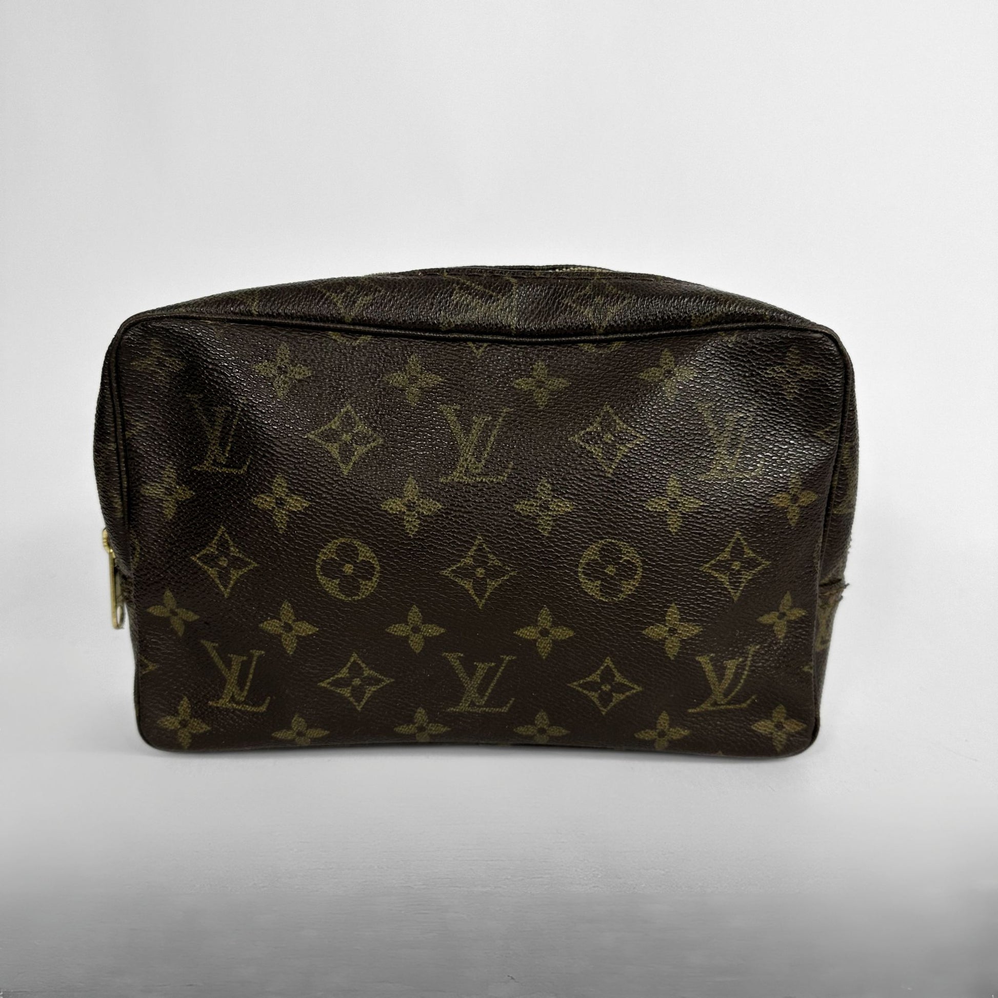 A Louis Vuitton monogram toiletry bag in dark brown coated canvas with gold-tone hardware, photographed against a neutral light gray background.