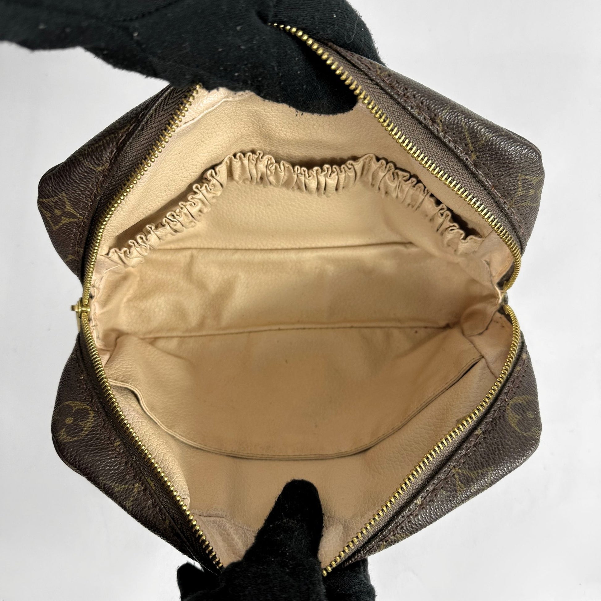 An open Louis Vuitton monogram canvas pouch showing the beige satin-lined interior, held open by black gloves against a plain light background.