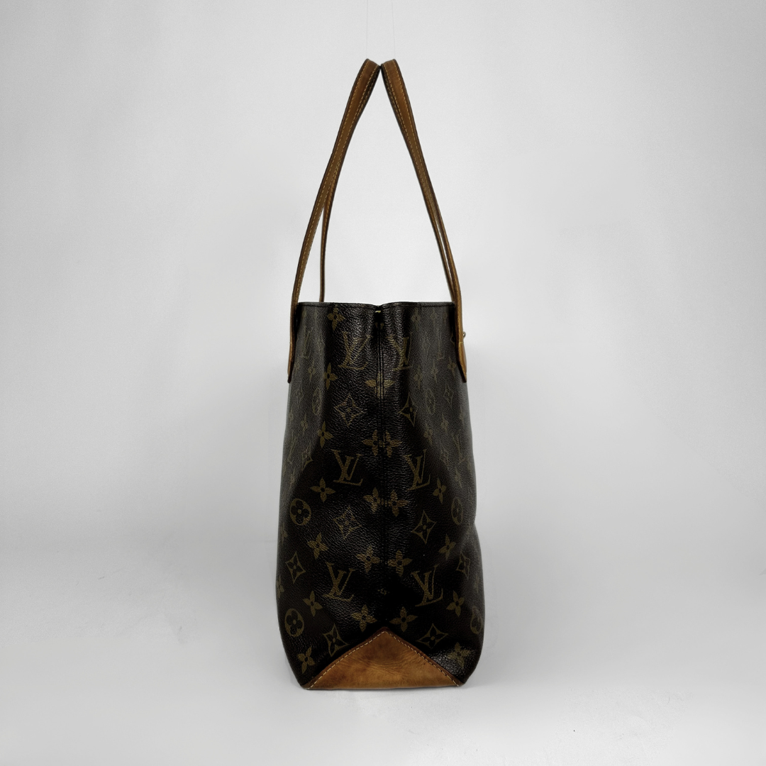 A Louis Vuitton monogram canvas tote bag in dark brown with tan leather shoulder straps and a tan leather bottom corner, shown against a light gray studio background.