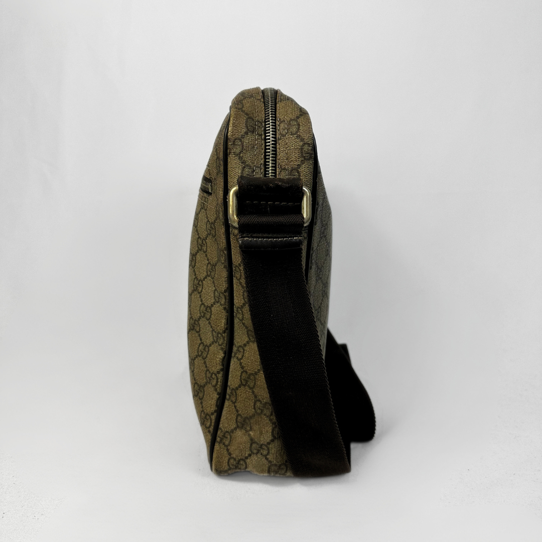 A Gucci monogram shoulder bag in brown and beige canvas with a black strap and gold-tone buckle, shown from a side profile view against a white background.