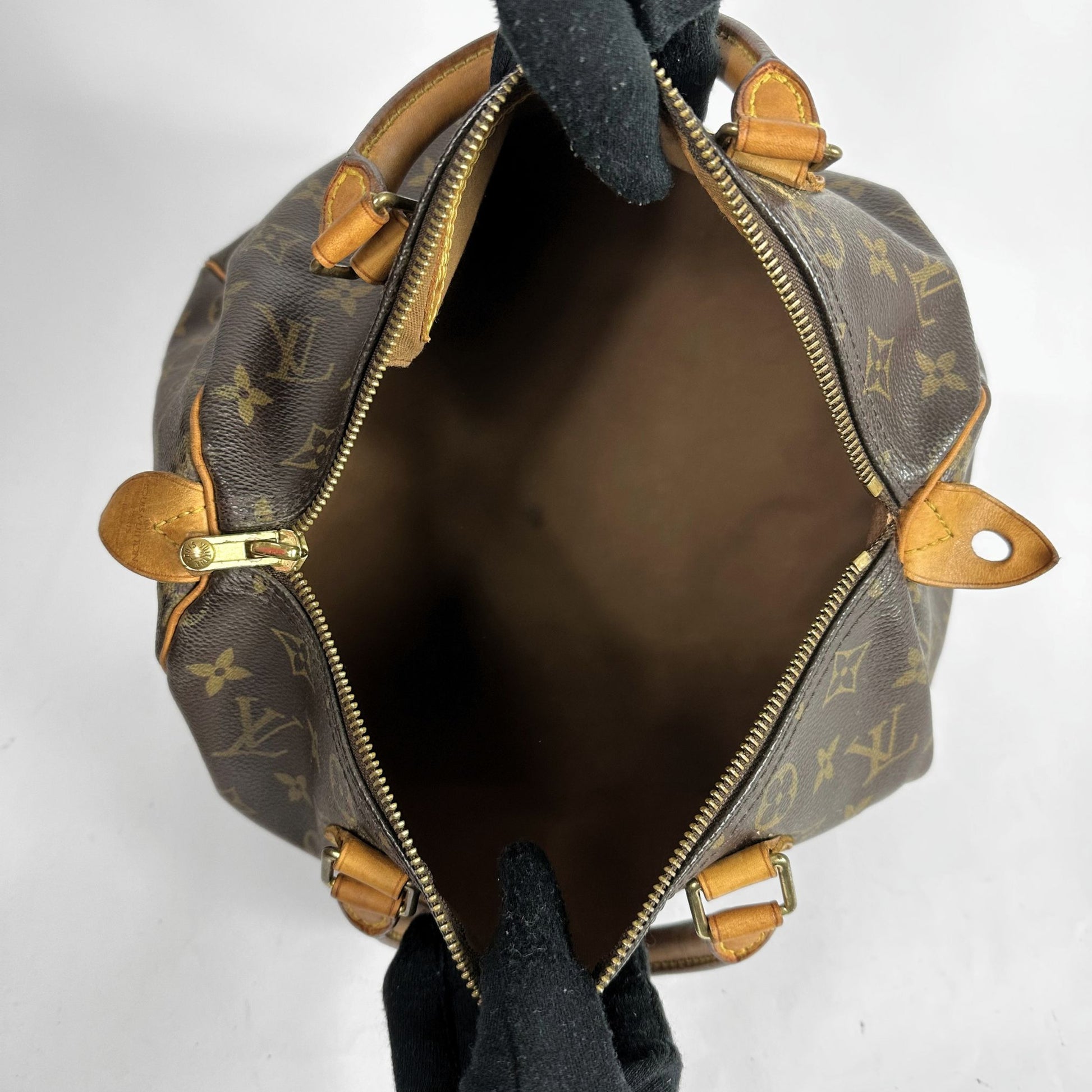 Louis Vuitton monogram canvas handbag shown open, viewed from the inside, with tan leather trim, gold-tone hardware, and black gloves holding the bag open against a plain light background.