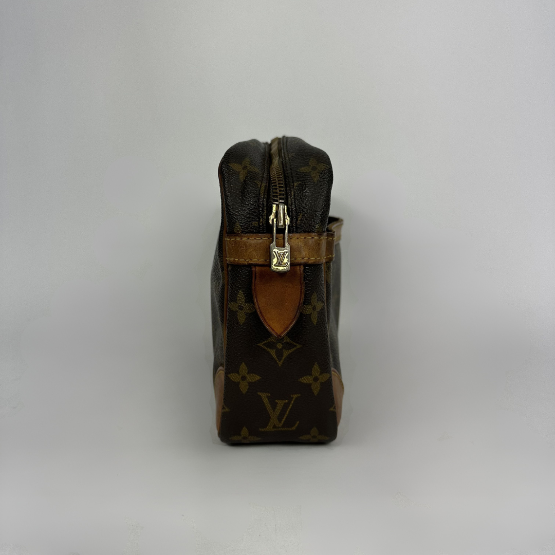 Louis Vuitton monogram canvas toiletry bag with brown leather trim and gold-tone zipper hardware, photographed against a neutral light gray background.