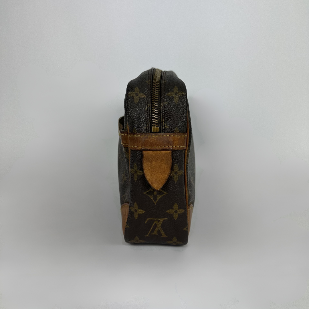 Louis Vuitton monogram canvas toiletry bag shown from the side, featuring dark brown coated canvas with tan monogram motifs and a tan leather strap detail, photographed against a neutral light gray background.