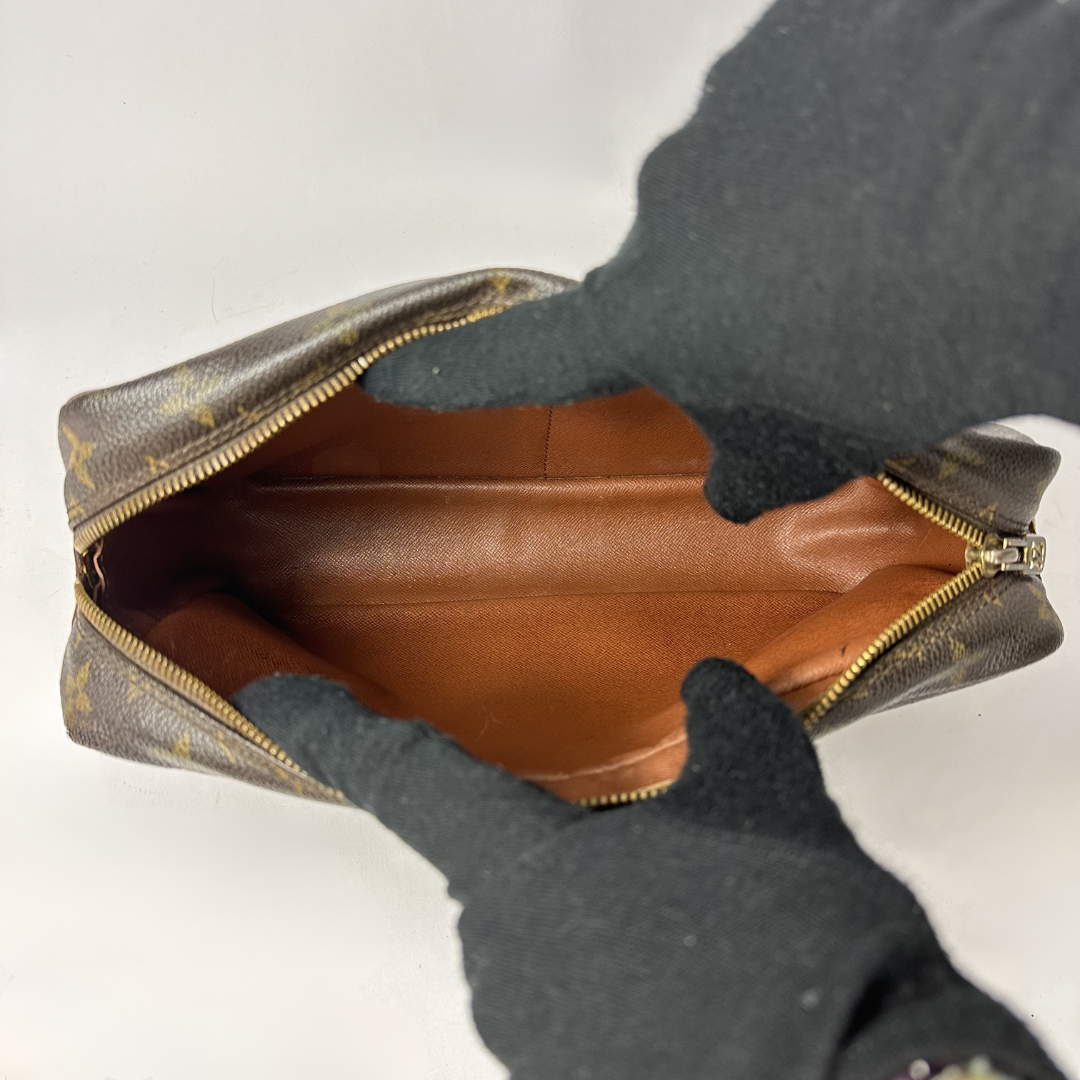A Louis Vuitton monogram canvas pouch shown open, revealing a brown leather interior, with a person wearing black gloves holding it against a plain white background.