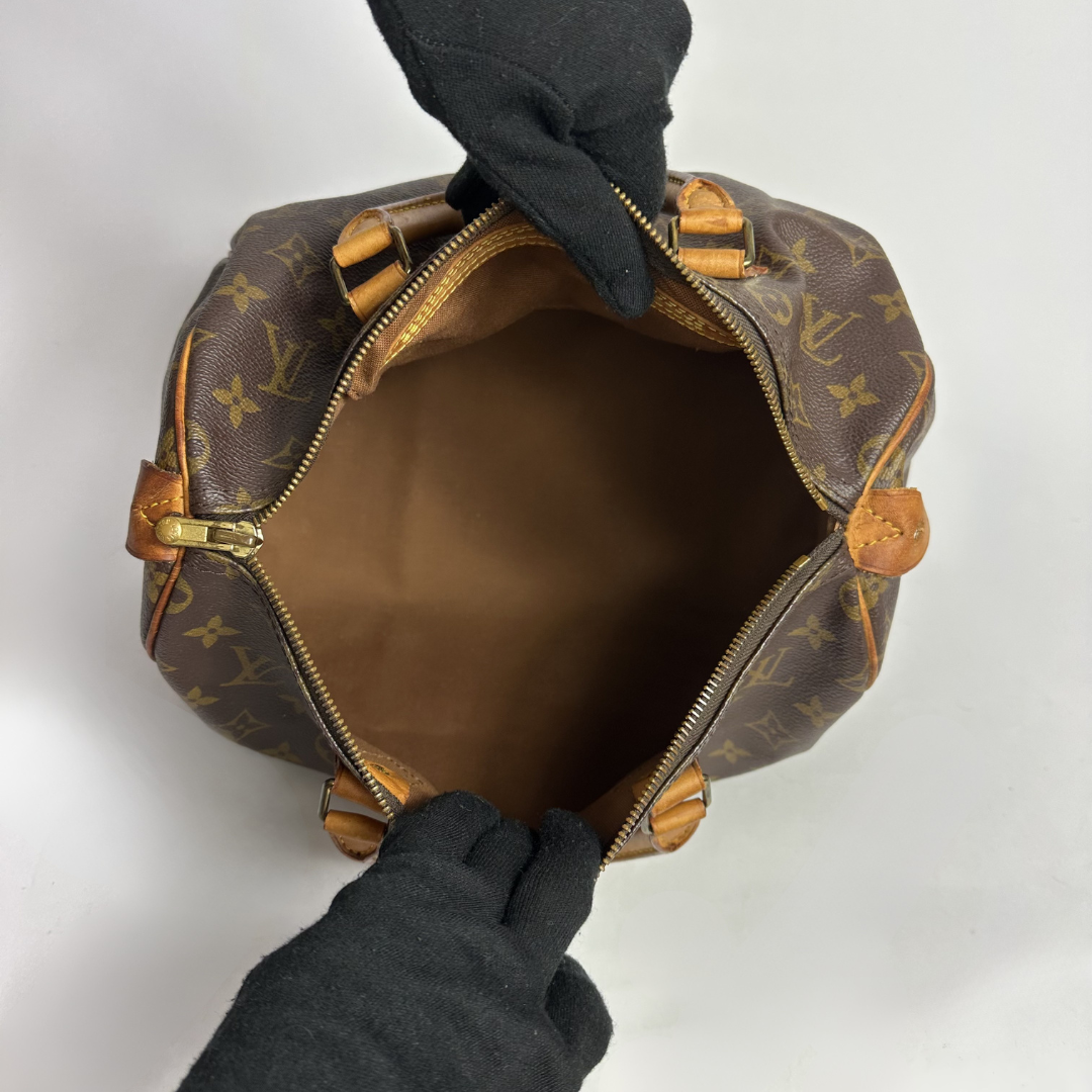 A Louis Vuitton monogram canvas handbag shown open from above, with a brown interior and gold-tone hardware. Two black-gloved hands are holding the bag open against a plain light background.