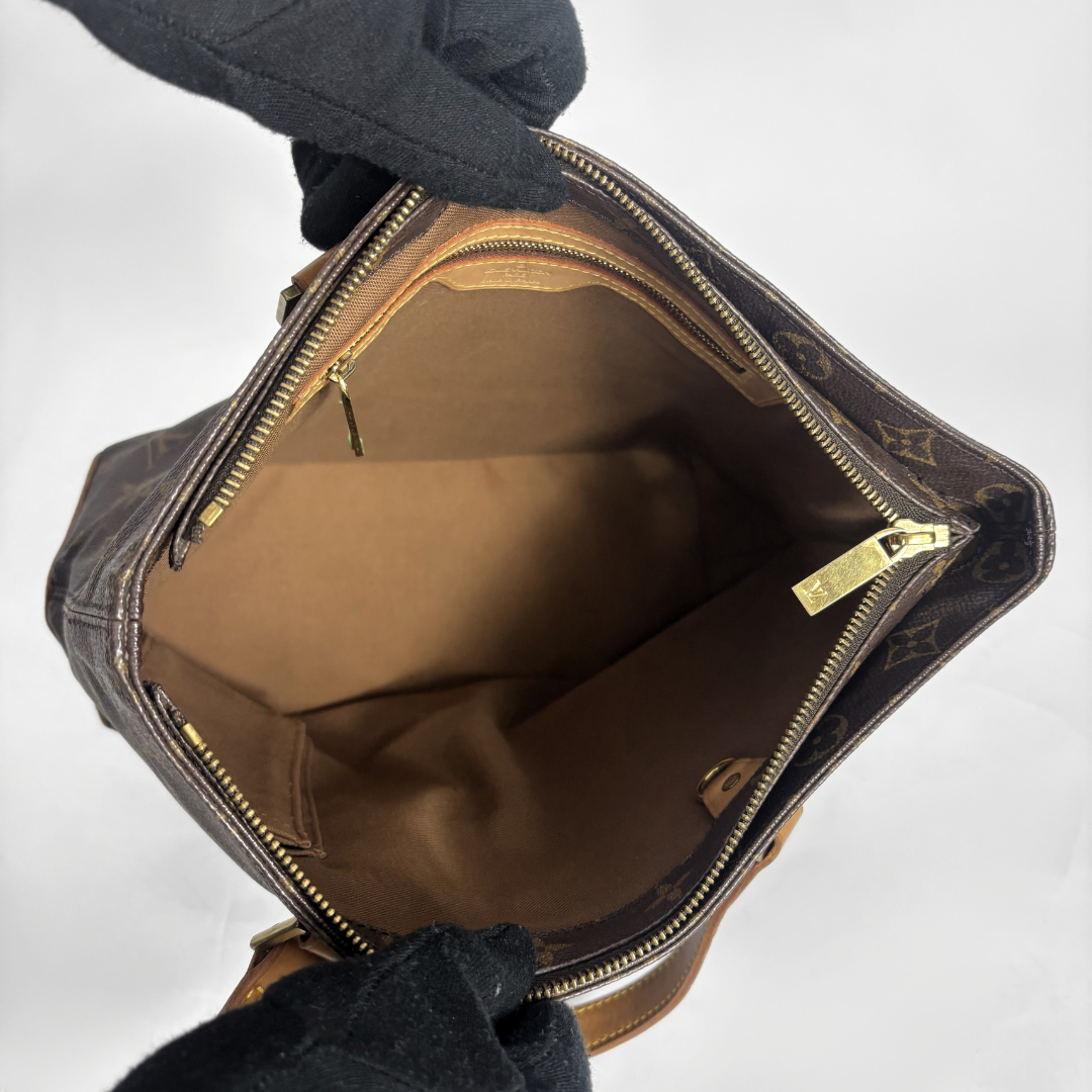 A Louis Vuitton monogram canvas handbag shown open, revealing a tan interior lining and gold-tone zippers, with a black gloved hand holding it against a plain white background.