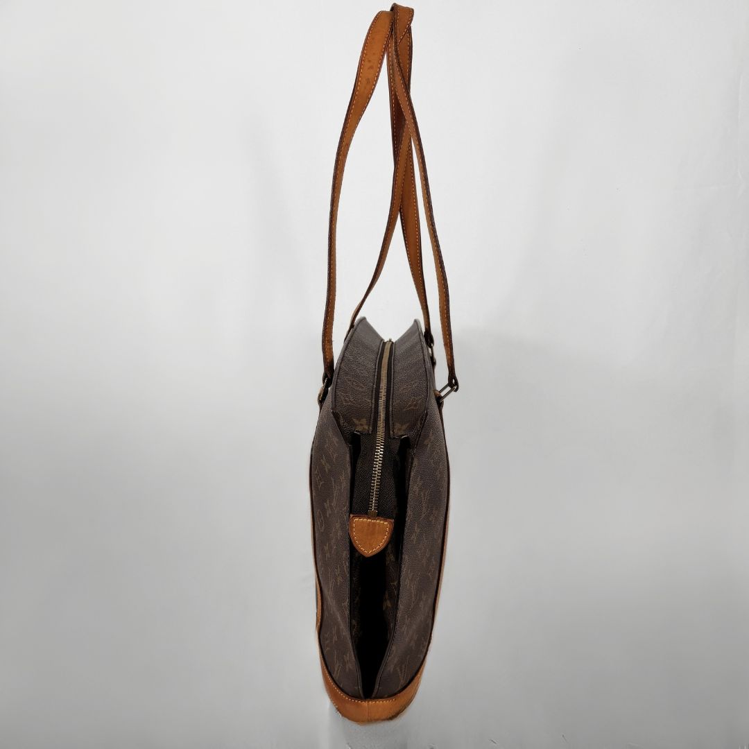 A brown monogram canvas shoulder bag with tan leather trim and strap, shown from a top-down view on a light gray background.