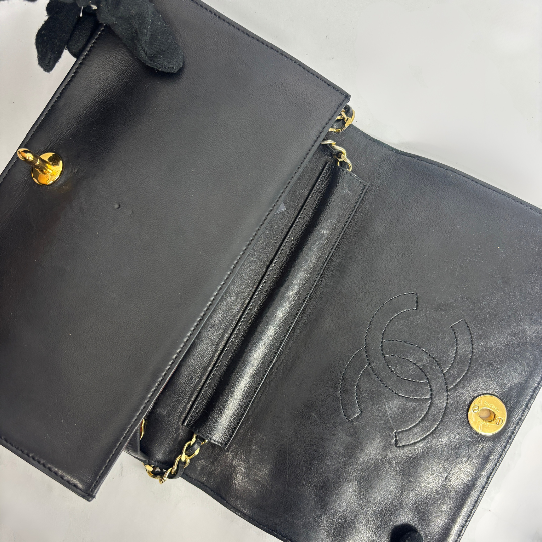 A black leather clutch bag with gold-tone hardware, shown open to reveal the interior. The right side features an embossed interlocking CC logo, characteristic of Chanel.