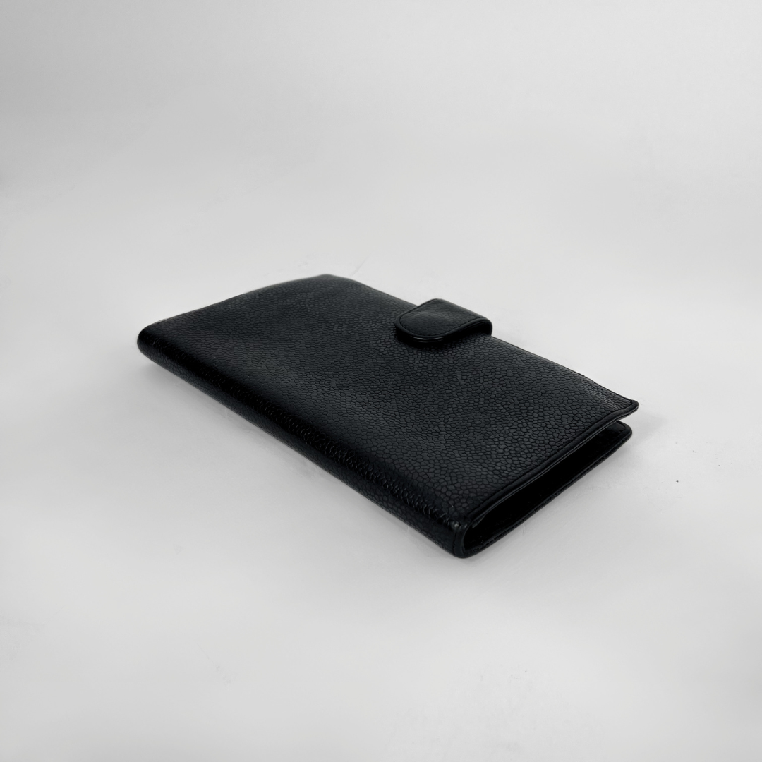 A black textured leather wallet with a small tab closure on the front, photographed on a light gray studio background.