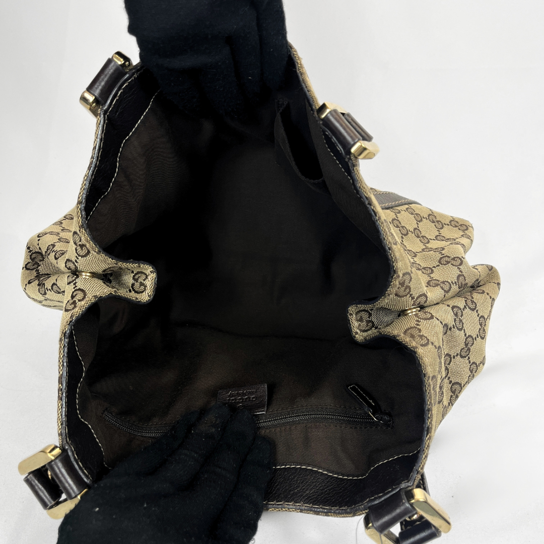 An open Gucci shoulder bag with black leather trim and gold-tone hardware, shown from the inside. The bag features the signature beige and brown GG monogram canvas, a black interior lining, and is held open by black gloves against a plain white background.