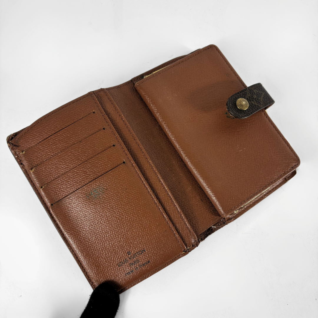 A brown textured leather bi-fold wallet shown open, revealing multiple card slots on one side and a small pocket with a snap-button strap on the other. A black gloved hand is visible holding the bottom edge against a plain white background.