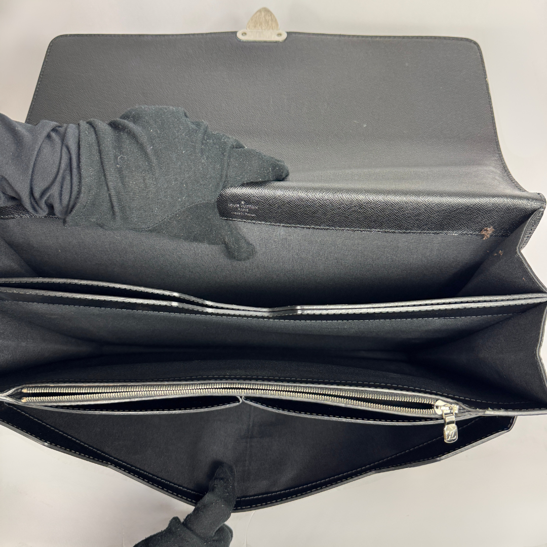 A black leather clutch or document holder shown open, with interior compartments and silver-tone zippers visible. 