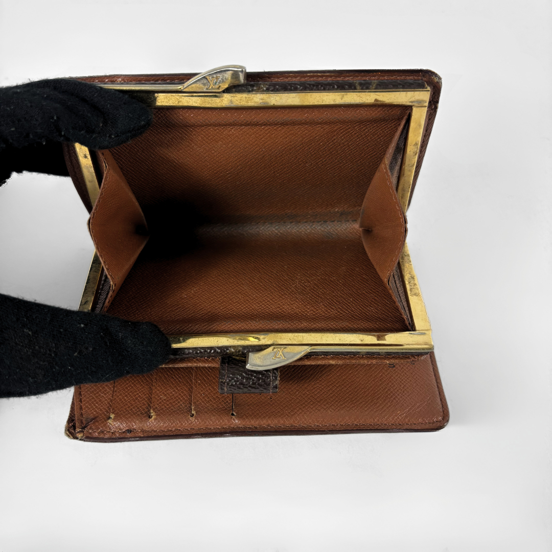 A vintage brown leather wallet or clutch with gold-tone metal frame and clasp, held open by a black gloved hand against a white background.
