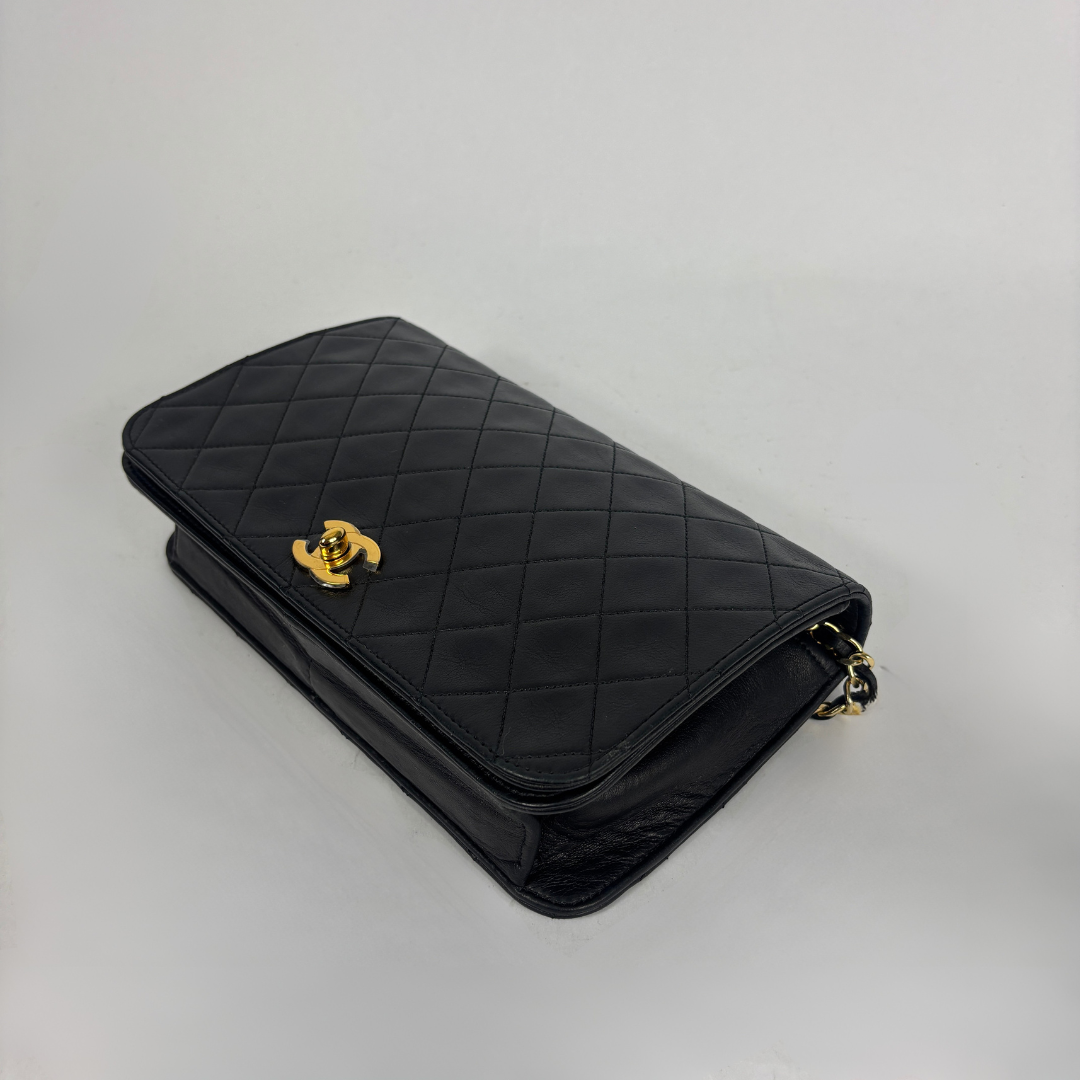 A black quilted leather shoulder bag with a gold-tone interlocking CC clasp, characteristic of Chanel, shown on a neutral light gray background.