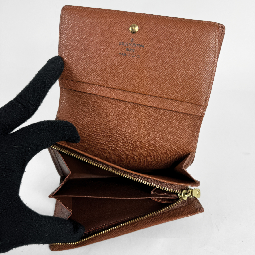 A brown textured leather wallet held open by a black gloved hand, showing interior card slots and a gold-tone zipper on a plain light background.