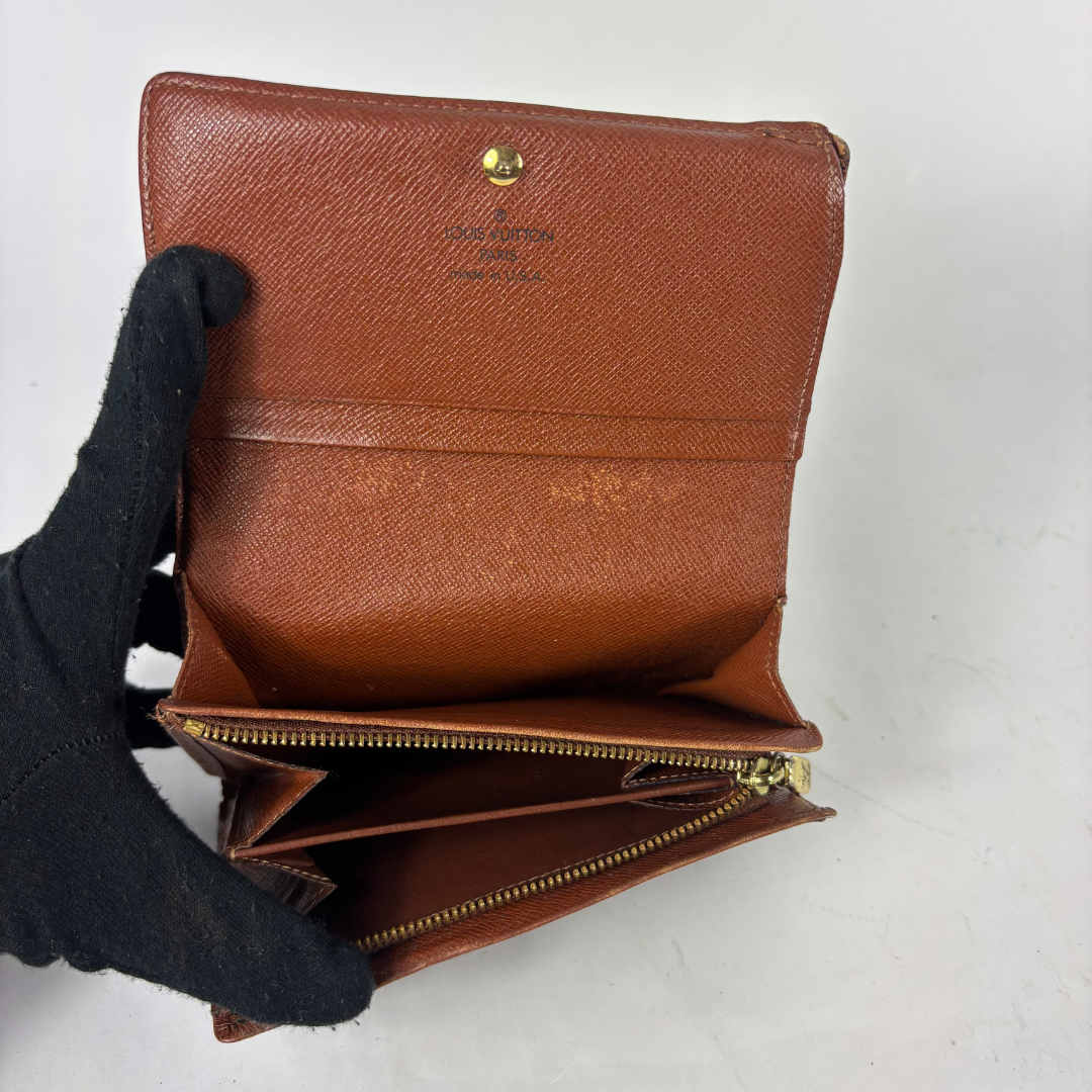 A brown textured leather wallet held open by a black gloved hand, showing its interior compartments and gold-tone zipper against a plain light background.